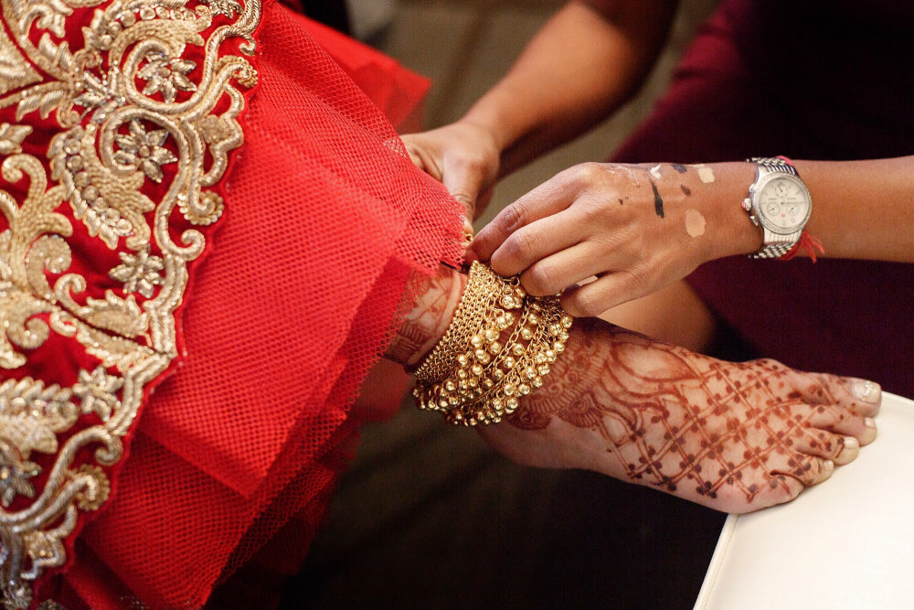 Gold bridal bangles being placed on a bride's henna-adorned wrist against a red embroidered lehenga